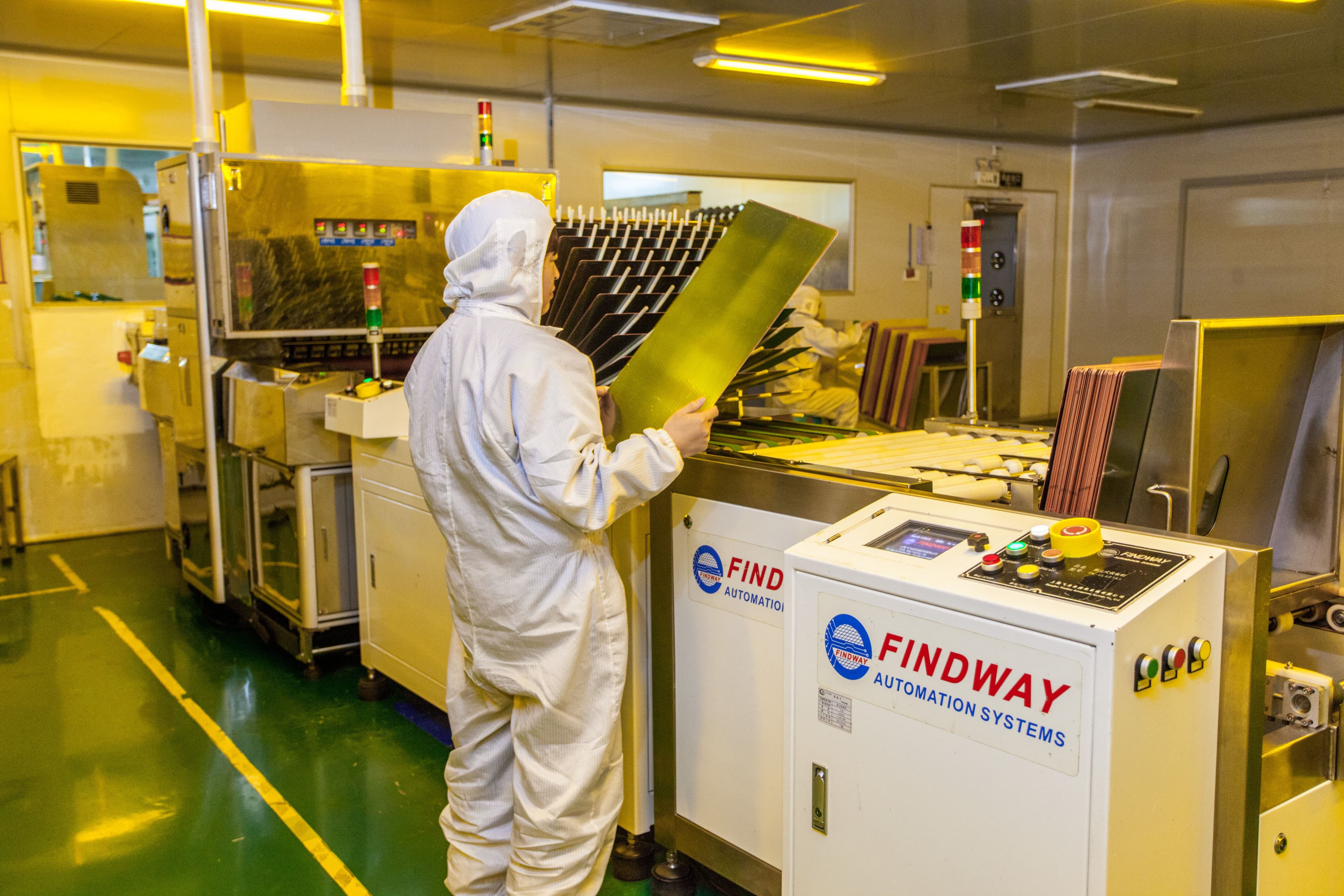 Technician handling a PCB panel beside automated PCB processing equipment.